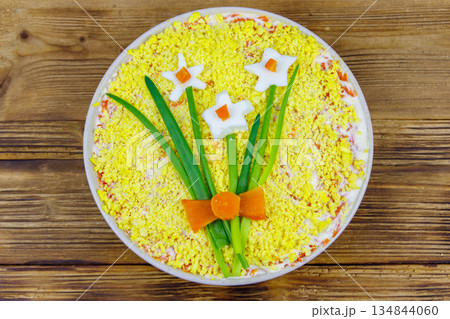 Traditional russian layered salad Mimosa with spring decoration Daffodil on wooden table. Top view. Decoration is made of egg, green onion and carrot 134844060