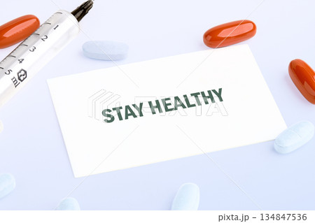 Assortment of orange and white pills, alongside a syringe, frame a card that reads STAY HEALTHY. They rest against a light-colored background Assortment of orange and white pills, alongside a syringe, frame a card that reads STAY HEALTHY. They rest against a light-colored background 134847536