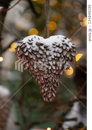 Handmade heart-shaped Christmas ornament made of natural seeds hangs on branches, covered with light snow, warm golden bokeh lights in the background Handmade heart-shaped Christmas ornament made of natural seeds hangs on branches, covered with light snow, warm golden bokeh lights in the background 134849195