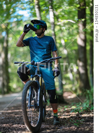 Man on mountain bike on forest trail drinks water during ride in sunny woods 134850131