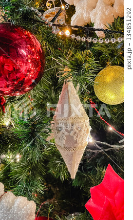 Close-up of an elegant cream textile ornament with lace details hanging on a Christmas tree with colorful baubles and lights. 134851292