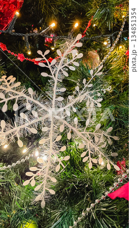 Close-up of a large white decorative snowflake hanging on a Christmas tree with red ornaments. Close-up of a large white decorative snowflake hanging on a Christmas tree with red ornaments. 134851354