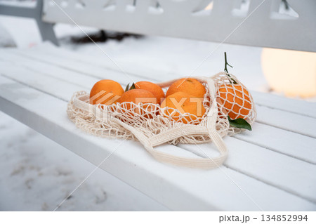 Fresh mandarins lie in a reusable mesh bag on a white bench with snow, creating a clean winter still life with bright orange accents Fresh mandarins lie in a reusable mesh bag on a white bench with snow, creating a clean winter still life with bright orange accents 134852994