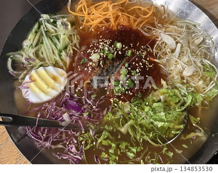 Chilled, chewy naengmyeon noodles in tangy broth paired with juicy, savory mandu dumplings, offering a perfect balance of refreshing coolness and hearty comfort in every delicious Korean bite. 134853530