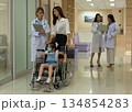 Medical team caring for girl patient in wheelchair at modern hospital. Doctor and mother accompany child wearing face mask through healthcare facility corridor with professional staff 134854283