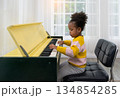 Young girl sitting on black cushioned chair playing piano in bright room with large window 134854285