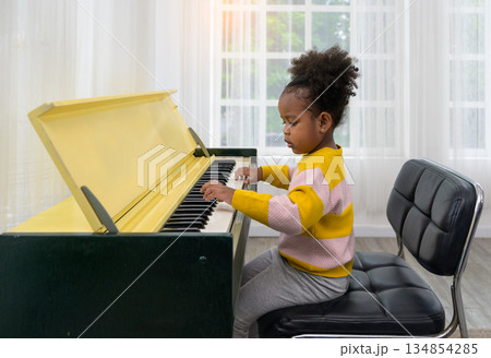 Young girl sitting on black cushioned chair playing piano in bright room with large window 134854285