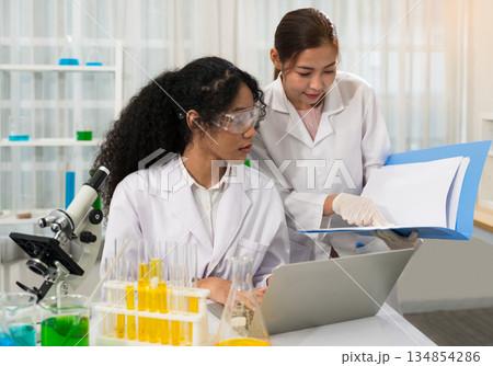 Two female scientists collaborate in laboratory with one wearing safety goggles while working on microscope and laptop with colorful test tubes. Other woman standing and reviewing research documents 134854286