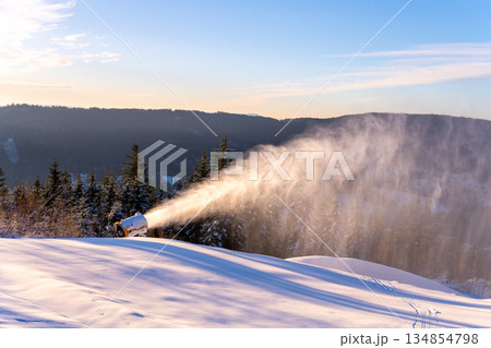 Snow cannon operates on a ski slope to produce artificial snow. The sun shines over the mountain landscape while snow accumulates for winter sports activities. 134854798