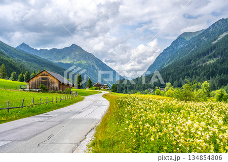 A winding road is seen under cloudy skies surrounded by mountains and green fields. Wooden buildings are scattered in the valley, showing rural life in the Alps. A winding road is seen under cloudy skies surrounded by mountains and green fields. Wooden buildings are scattered in the valley, showing rural life in the Alps. 134854806