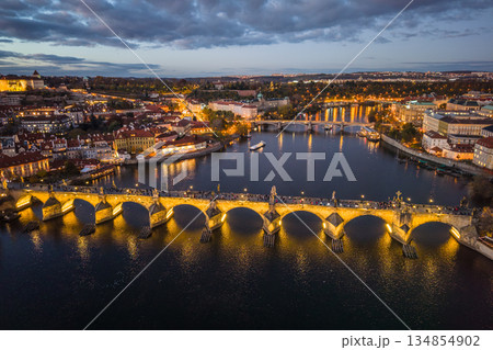 Aerial view of Charles Bridge illuminated at night. The scene showcases the water below, surrounding buildings, and the vibrant lights of Prague. Evening time highlights the beauty of this location. Aerial view of Charles Bridge illuminated at night. The scene showcases the water below, surrounding buildings, and the vibrant lights of Prague. Evening time highlights the beauty of this location. 134854902