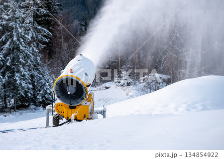 A snow cannon creates artificial snow on a winter slope. The machine blows snow into the air, covering the ground. Nearby trees are dusted with white snow in a ski area. A snow cannon creates artificial snow on a winter slope. The machine blows snow into the air, covering the ground. Nearby trees are dusted with white snow in a ski area. 134854922