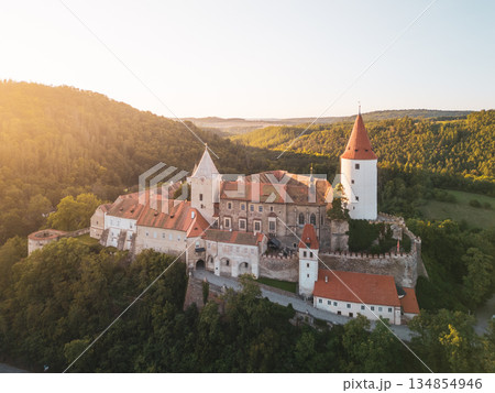 Krivoklat Castle stands proudly atop a hillside bathed in golden sunset light. Surrounded by lush greenery, this medieval structure reflects its rich history. The serene landscape adds to its charm. 134854946