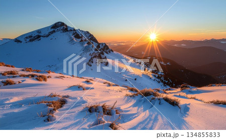 Sunrise illuminates a snow-covered mountain range. The rising sun paints the snow-covered landscape with warm light. Dramatic lighting highlights the mountainous terrain and distant peaks. 134855833