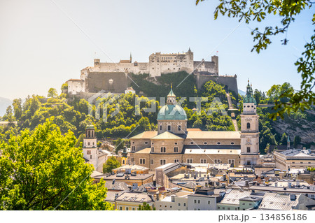 Salzburg showcases its majestic castle perched on the hill, surrounded by lush greenery. The city below features charming buildings and a beautiful river, creating a stunning view. 134856186