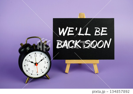 We'll Be Back Soon text with easel and alarm clock on purple background We'll Be Back Soon text with easel and alarm clock on purple background 134857892