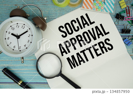 Social Approval Matters text on a paper surrounded by office supplies, a magnifying glass, and a clock on a blue wooden surface 134857950