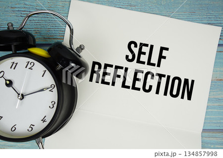 Notepad on a blue wooden table with the words "Self Reflection" and alarm clock 134857998