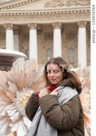 Woman stands near a snow-covered fountain with large white flower decorations in front of the Bolshoi Theatre facade and colonnade 134858364