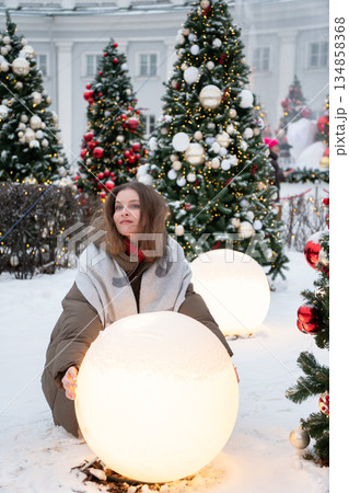 Woman in winter clothing touches glowing spherical light installation among decorated Christmas trees, snow, festive lights in modern holiday setting 134858368