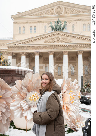 Woman stands near a snow-covered fountain with large white flower decorations in front of the Bolshoi Theatre facade and colonnade 134858369