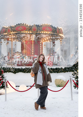Smiling woman poses near glowing carousel, decorated Christmas trees, holding reusable mesh bag with mandarins in snowy festive setting 134858390