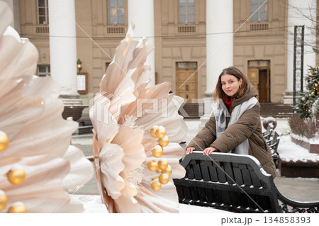Woman stands by a bench near a large floral New Year installation in front of the Bolshoi Theatre, with snow-covered square and classical columns 134858393