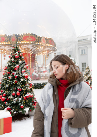 Woman in winter jacket and scarf stands near decorated Christmas trees and glowing carousel, touching her hair amid snow and warm festive lights 134858394