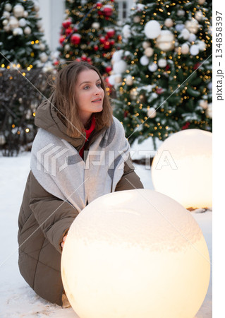 Woman in winter clothing touches glowing spherical light installation among decorated Christmas trees, snow, festive lights in modern holiday setting Woman in winter clothing touches glowing spherical light installation among decorated Christmas trees, snow, festive lights in modern holiday setting 134858397