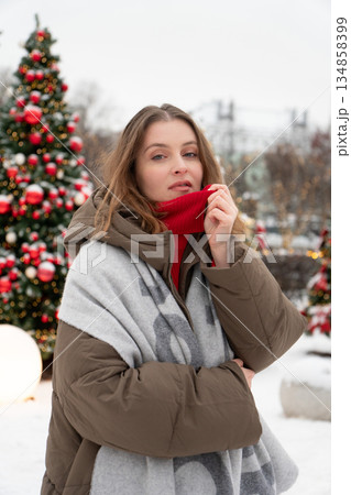 Young woman in warm winter jacket and scarf stands outdoors near decorated Christmas trees with red ornaments, snow, and soft festive lights Young woman in warm winter jacket and scarf stands outdoors near decorated Christmas trees with red ornaments, snow, and soft festive lights 134858399