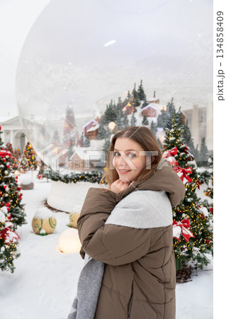 Smiling woman in warm winter jacket stands among decorated Christmas trees with red ornaments in a snowy Nutcracker-themed New Year installation Smiling woman in warm winter jacket stands among decorated Christmas trees with red ornaments in a snowy Nutcracker-themed New Year installation 134858409