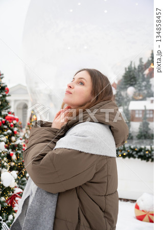 Smiling woman in warm winter jacket stands among decorated Christmas trees with red ornaments in a snowy Nutcracker-themed New Year installation Smiling woman in warm winter jacket stands among decorated Christmas trees with red ornaments in a snowy Nutcracker-themed New Year installation 134858457