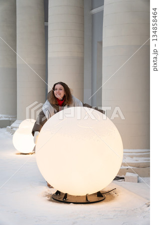 Smiling woman in winter clothing interacts with glowing spherical light installations in a snowy urban space, creating a modern festive winter mood 134858464