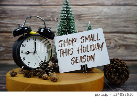 Festive holiday scene featuring a vintage alarm clock, pinecones, and miniature Christmas trees on a wooden base, with the text "Shop Small This Holiday Season" encouraging support for local businesse 134858610