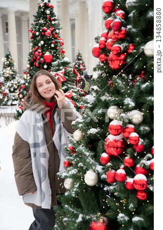 Woman in winter jacket stands beside decorated Christmas trees with red, white ornaments, snow, warm lights, creating cozy festive winter mood 134858880