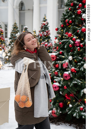 Playful woman in winter clothing holds mandarin and reusable mesh bag, posing among decorated Christmas trees with snow, festive lights Playful woman in winter clothing holds mandarin and reusable mesh bag, posing among decorated Christmas trees with snow, festive lights 134858889