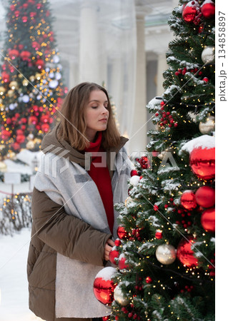 Woman in winter clothing stands close to decorated Christmas tree with red ornaments, snow, and warm festive lights, creating cozy holiday atmosphere Woman in winter clothing stands close to decorated Christmas tree with red ornaments, snow, and warm festive lights, creating cozy holiday atmosphere 134858897