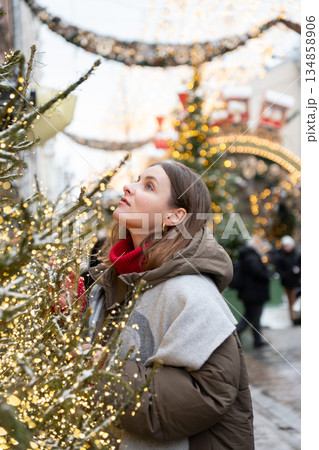 Woman in winter coat, red sweater looks up at glowing festive decoration among snow-dusted fir branches, surrounded by golden lights, holiday mood 134858906