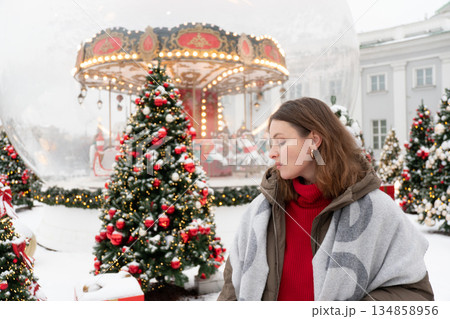 Woman in winter jacket and scarf stands near decorated Christmas trees and glowing carousel, touching her hair amid snow and warm festive lights Woman in winter jacket and scarf stands near decorated Christmas trees and glowing carousel, touching her hair amid snow and warm festive lights 134858956