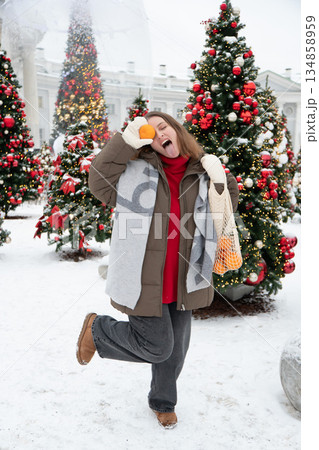 Playful woman in winter clothing holds mandarin and reusable mesh bag, posing among decorated Christmas trees with snow, festive lights Playful woman in winter clothing holds mandarin and reusable mesh bag, posing among decorated Christmas trees with snow, festive lights 134858959