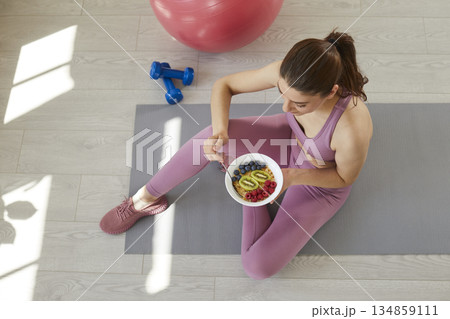Young woman sitting on workout mat and eating healthy cereal with fruit and berries Young woman sitting on workout mat and eating healthy cereal with fruit and berries 134859111