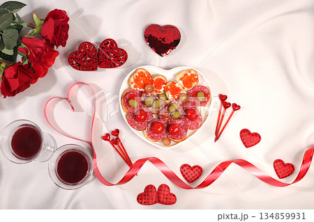 Heart of fresh canapes with salami, caviar, cheese and grapes for a buffet with roses, red wine and red hearts on the festive table. Valentine's Day dishes, advertising banner, bar invitations, menu 134859931
