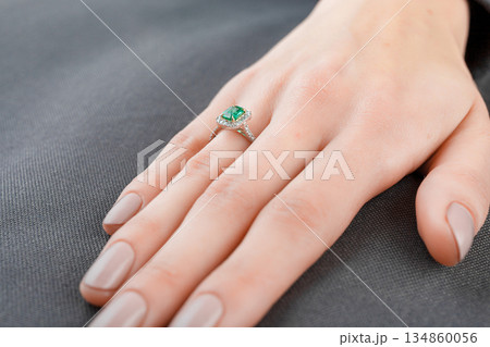 Beautiful female hand with gold ring with diamonds and emerald on gray background Beautiful female hand with gold ring with diamonds and emerald on gray background 134860056