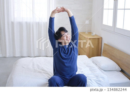 ベッドに座って伸びをする20代男性 134862244