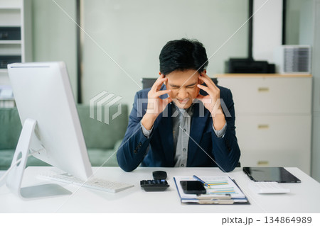 Frustrated young businessman working on a laptop computer sitting at his working place Frustrated young businessman working on a laptop computer sitting at his working place 134864989