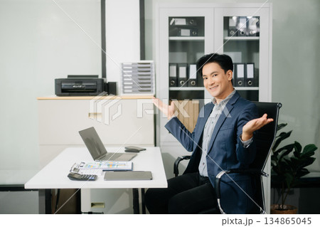 Confident businessman in suit, working on laptop and smartphone at office desk. 134865045