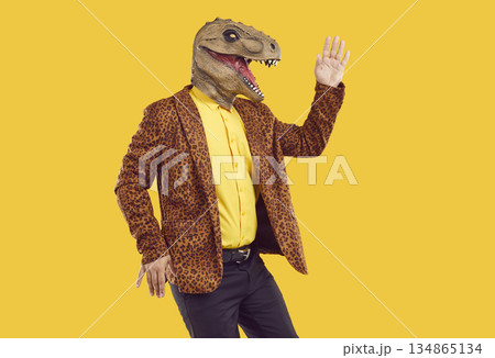 Eccentric funny showman in rubber dinosaur mask dancing and waving hand on yellow background. 134865134