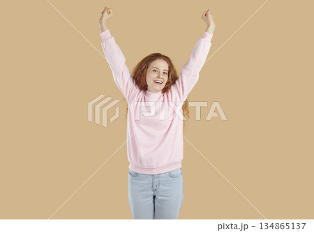 Curly redhead happy girl is rising her arms up on beige background and looking at camera. Curly redhead happy girl is rising her arms up on beige background and looking at camera. 134865137