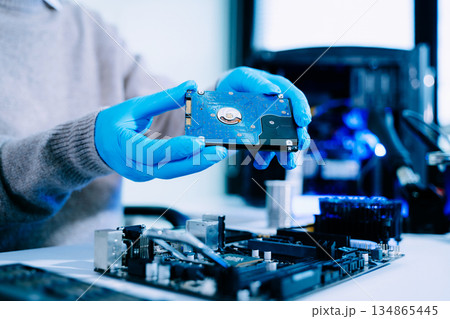 Closeup of technician with gloves holding blue hard drive circuit board above motherboard, symbolizing data recovery, storage, and computer repair. Closeup of technician with gloves holding blue hard drive circuit board above motherboard, symbolizing data recovery, storage, and computer repair. 134865445