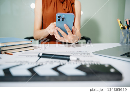 Creative woman multitasking with phone, clapperboard, and laptop while planning film production in a modern studio workspace. Behind the scenes workflow concept. 134865639
